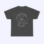The Thank God I'm Gay Shirt is dark gray and features a line drawing of a teddy bear surrounded by hearts and flowers, with “THANK GOD I’M GAY” in bold, playful letters above and below the bear.