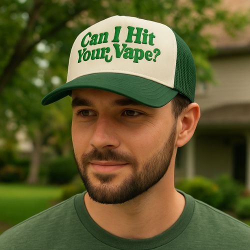 A man with a beard, wearing a green and white hat that says Can I Hit Your Vape? in green letters, stands outdoors with greenery and a house in the background.