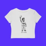 The Born Ready Shirt is a white crop top with a black-and-white illustration of a child in a baseball uniform, the words "Born Ready" below, and a solid bright blue background that makes it stand out.