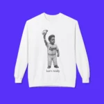 The Born Ready Shirt is a white tee showcasing a black-and-white illustration of a child in an Expos baseball uniform raising a cap, with the text "Born ready" beneath, all set against a bright blue background.