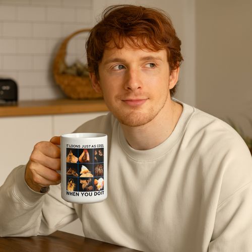 A young man with reddish hair, wearing a white sweatshirt, sits at a table holding a white mug with a meme featuring several pictures of cats. He is looking slightly to the side and smiling softly.