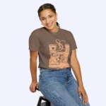 A young woman with braided hair, wearing the Sabrina Carpenter House Tour Shirt and blue jeans, sits on a black stool and smiles at the camera in front of a light grey background.